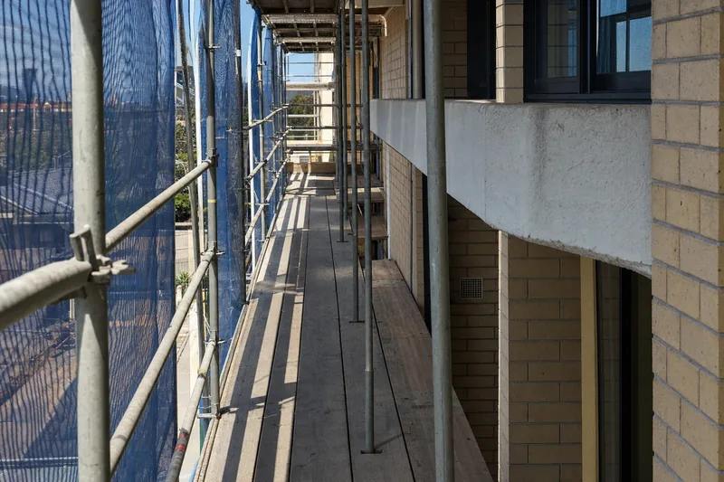 Scaffold and debris netting erected against one elevation of a blond-brick strata block, working decks at balcony level — illustrative example