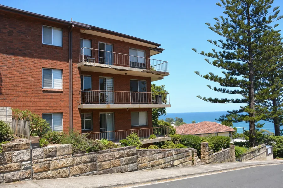 A three-storey liver-brick walk-up on a steep Eastern Suburbs street, stacked concrete balconies with render blown away at one slab edge, sandstone retaining wall and a Norfolk pine, ocean beyond — illustrative example