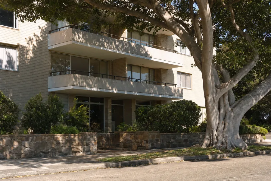 A cream-brick 1960s unit block under a mature Moreton Bay fig with stacked concrete balconies and a sandstone retaining wall, in soft harbour light — illustrative example