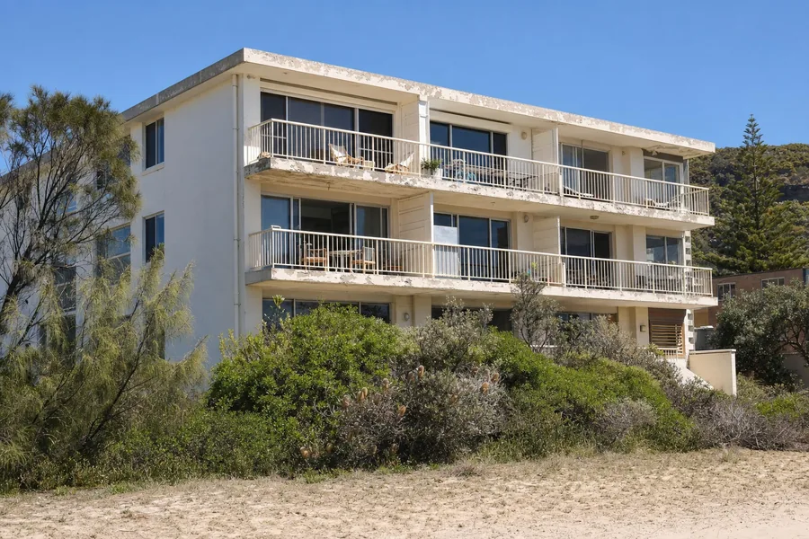 A low-rise rendered beachfront apartment block on the Northern Beaches with coating chalked and lifting along every concrete balcony edge, banksia in the foreground and the escarpment behind — illustrative example