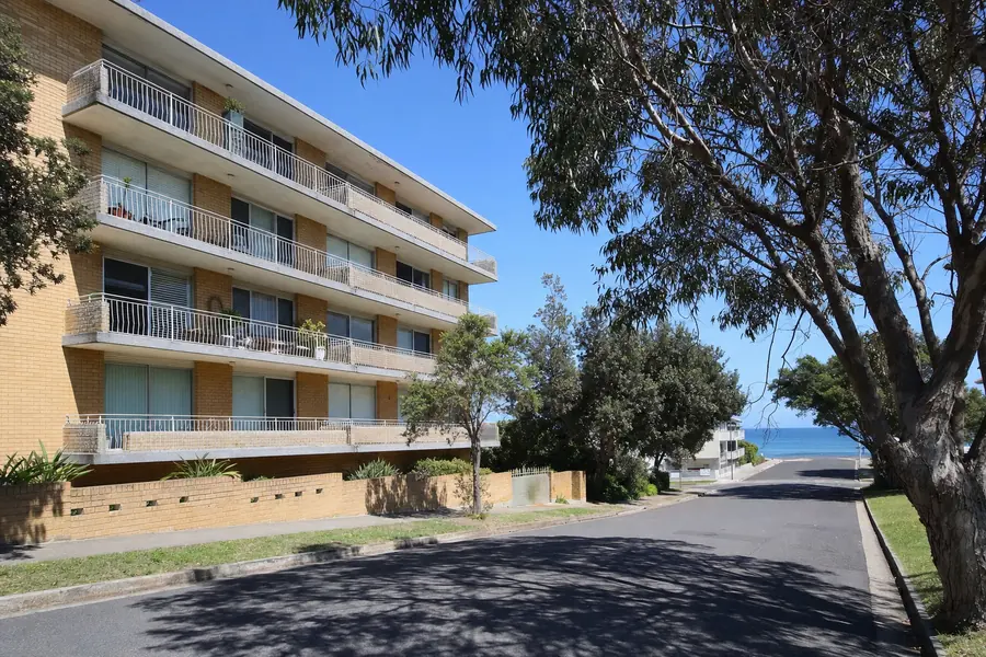 A four-storey blond-brick strata block on a wide quiet street near Cronulla with deep concrete balcony slabs, a gum in the verge and water visible at the end of the road — illustrative example