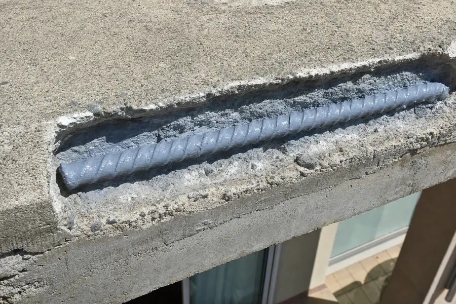 Exposed reinforcing bar in a concrete balcony edge, wire-brushed clean and coated with anti-corrosion primer, the recess prepared and empty before reinstatement — illustrative example